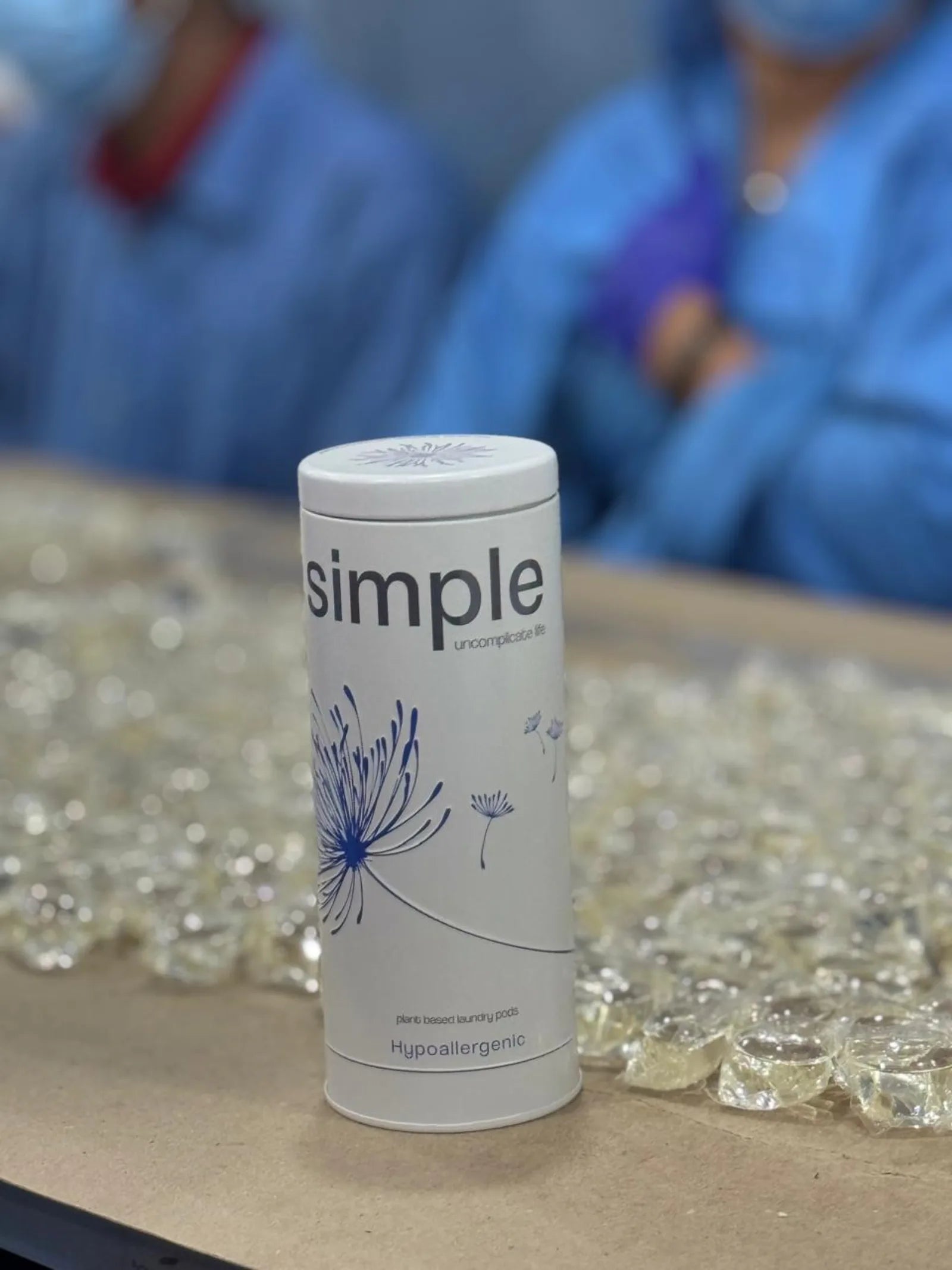 A simple Hypoallergenic tin on a tray beside loose clear pods on the production floor in India, with two workers in blue coats behind it